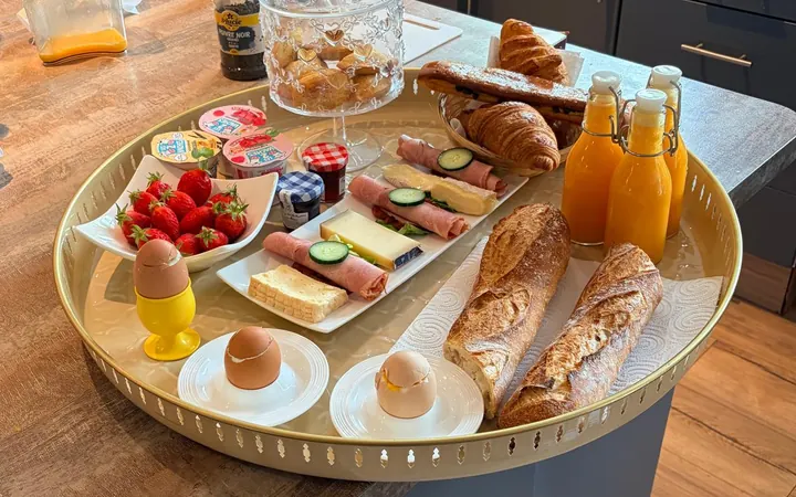 Un plateau doré sur le plan de travail de la cuisine : œufs à la coque, fraises, fromage et jambon, baguettes tradition, croissants et jus d’orange pressé