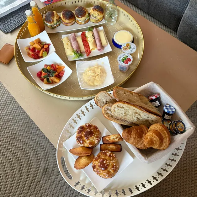 Deux plateaux sur la table : buns, fromage, jambon, salades de fruits, œufs brouillés et jus d’orange sur l’un, pain, croissants, brioches et madeleines sur l’autre