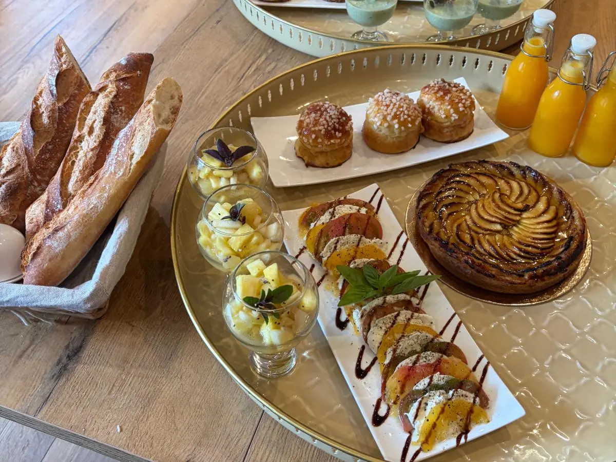 Les plateaux du matin dressés sur une table en bois : brioches au sucre, tarte aux pommes, tomates et mozzarella, salades d’ananas, baguettes et jus d’orange pressé