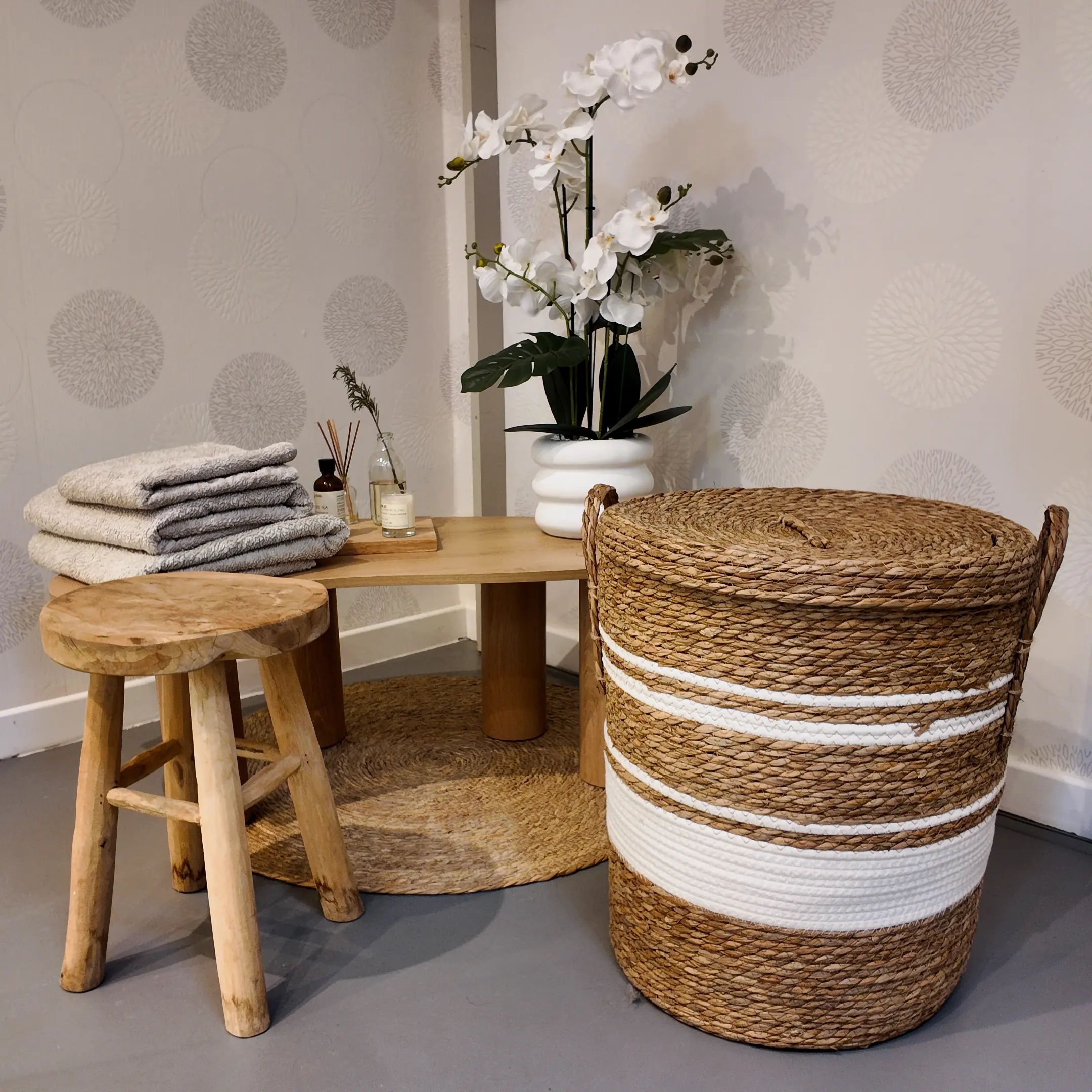 Des serviettes pliées sur une table basse, un tabouret de bois brut, une orchidée blanche et un grand panier tressé