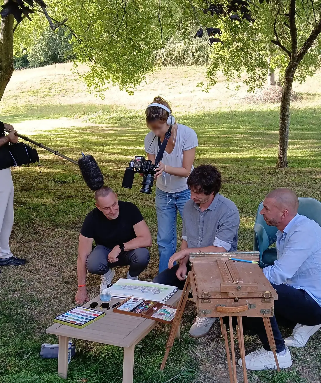 Le tournage au Hameau des Prés-Verts : Stéphane, accroupi, et Loïc, assis, penchés avec Cyp sur son aquarelle ; une cadreuse de France 2 filme au-dessus d’eux, une perche de son entre dans le cadre